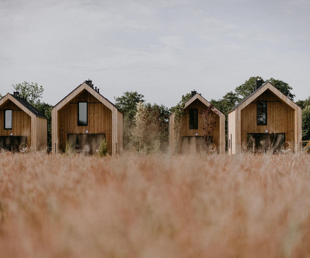 Cztery nowoczesne budynki rekreacji indywidualnej projektu Mariusza Głębockiego, z elewacjami z naturalnego drewna i grafitowymi dachami, usytuowane w sielskim otoczeniu wysokiej, suchej trawy i zielonych drzew. O projekcie przeczytasz na Architektura Murator Plus.