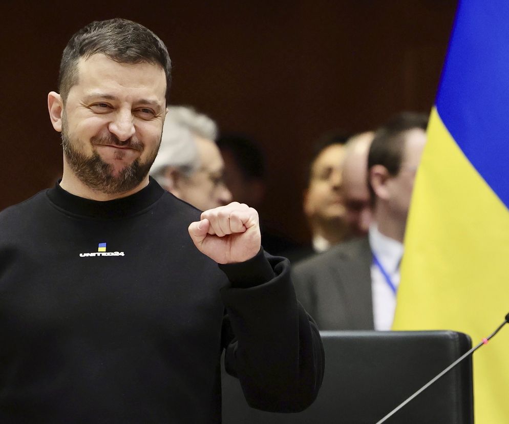 Prezydent Ukrainy Wołodymyr Zełenski z brodą, ubrany w czarny sweter z logo UNITED24, zaciśniętą pięścią symbolizuje determinację. W tle widoczna flaga Ukrainy. Zdjęcie wykonane w Brukseli, gdzie Zełenski omawiał wsparcie finansowe i postępy w walce z korupcją, o czym przeczytasz na Super Biznes.