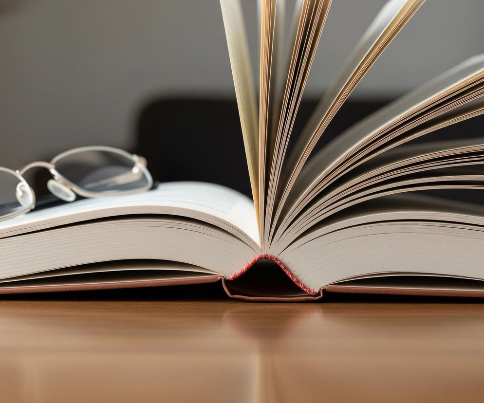 Otwarta książka leży na drewnianym blacie, z rozłożonymi stronami skierowanymi ku górze, a wiele z nich jest rozchylonych i widocznych od boku. Oprawka książki jest brązowa, a grzbiet, z widocznym czerwonym szwem, jest skierowany do widza. Na lewej stronie książki leżą srebrne okulary z przezroczystymi szkłami. Tło jest rozmyte, z widocznymi odcieniami szarości i czerni.