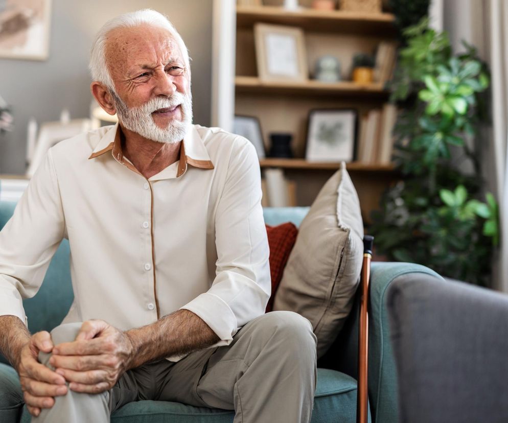 Starszy mężczyzna z siwą brodą, siedzący na turkusowej kanapie, trzyma dłonie na bolącym kolanie, wyrażając grymasem cierpienie. Obraz ilustruje problem bólu stawów kolanowych, nasilający się przed deszczem. Więcej informacji na ten temat znajdziesz na Poradnik Zdrowie.