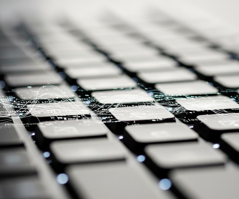 Na zdjęciu widoczna jest klawiatura laptopa w ujęciu z góry pod kątem, z perspektywą zbiegającą się w górnej części obrazu. Dominują ciemne klawisze o zaokrąglonych narożnikach i jasnych symbolach, częściowo rozmyte. Na górze klawiszy, zwłaszcza w lewej części, widoczne są białe, świecące linie i wykresy, tworzące skomplikowany wzór danych i połączeń, co nadaje obrazowi futurystyczny charakter. Głęboka ostrość koncentruje się na pierwszym planie i środku kadru, natomiast dalsze rzędy klawiszy są rozmazane, tworząc efekt bokeh. Całość utrzymana jest w ciemnej tonacji z jasnymi, niebieskawymi akcentami świateł i linii.