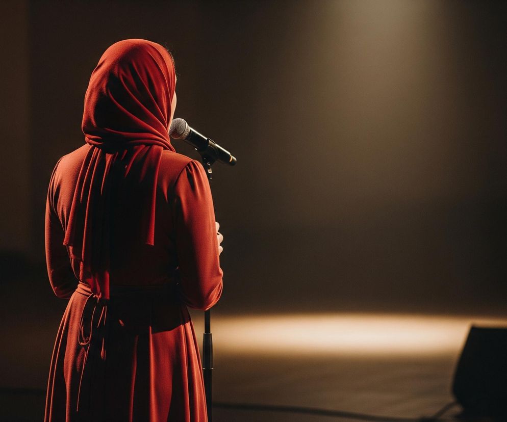 Kobieta stoi tyłem do obserwatora, zwrócona w stronę mikrofonu na statywie, który znajduje się przed nią. Ma na sobie długą, czerwoną sukienkę i hidżab w tym samym kolorze. Prawa ręka kobiety jest lekko uniesiona i znajduje się w pobliżu mikrofonu. Cała scena jest słabo oświetlona, z punktowym światłem padającym na kobietę i mikrofon z góry oraz lekką poświatą w tle.
