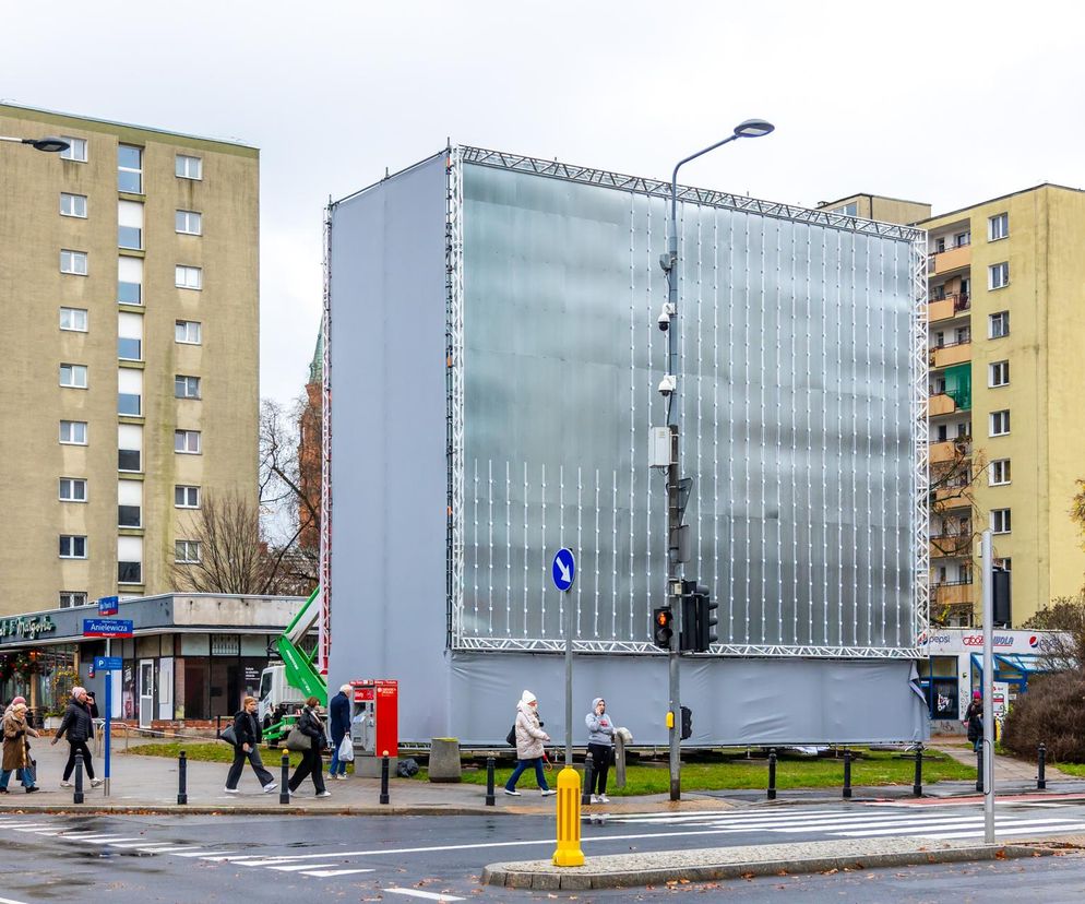 Ogromny, nielegalny stelaż reklamowy u zbiegu ulic Anielewicza i Jana Pawła II w Warszawie, zasłaniający kamienice i drzewa. Widoczni przechodnie oraz sygnalizacja świetlna. O nielegalnych reklamach w przestrzeni miejskiej przeczytasz na Architektura Murator Plus.