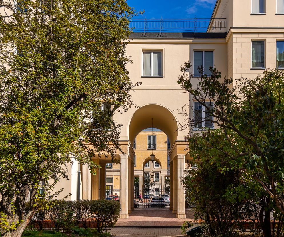 Ujęcie na arkadę prowadzącą do wewnętrznego dziedzińca Osiedla Kubusia Puchatka (Nowy Świat Zachód) w Warszawie. Kremowe elewacje, klasyczne kolumny i zieleń drzew podkreślają socrealistyczny styl. Architektura Murator Plus to portal, na którym można przeczytać o tym osiedlu.