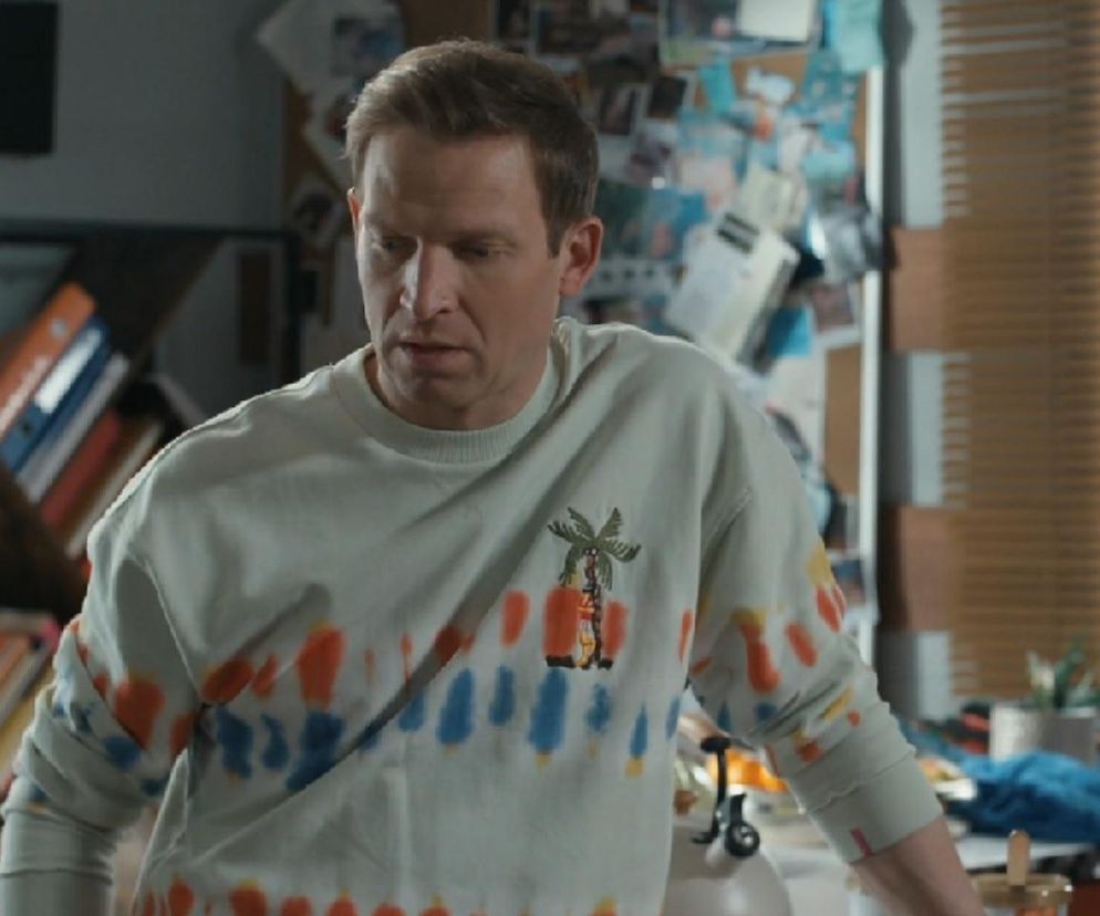 Michał Rolnicki jako Łukasz Sadowski w jasnej bluzie z kolorowymi plamami i naszywką palmy, zmartwiony problemami finansowymi, w scenie z serialu „Barwy Szczęścia” - więcej informacji o jego losach znajdziesz na portalu Super Seriale.