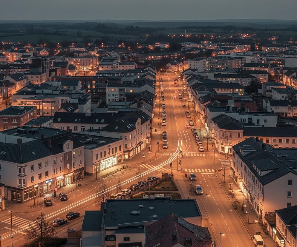 Miasto widoczne z lotu ptaka o zmierzchu lub wczesnym ranku, z licznymi budynkami o jasnych elewacjach i ciemnych dachach. Główna ulica przecina obraz od dołu do góry, rozświetlona pomarańczowym blaskiem latarni, z widocznymi smugami świateł pojazdów wskazującymi na ruch. Po obu stronach ulicy stoją rzędy budynków, a w oddali widać ciemne pola i zalesione wzgórza z rozsianymi punktami świateł. Panuje chłodna, niebieskawo-szara poświata nieba kontrastująca z ciepłymi barwami miejskiego oświetlenia.