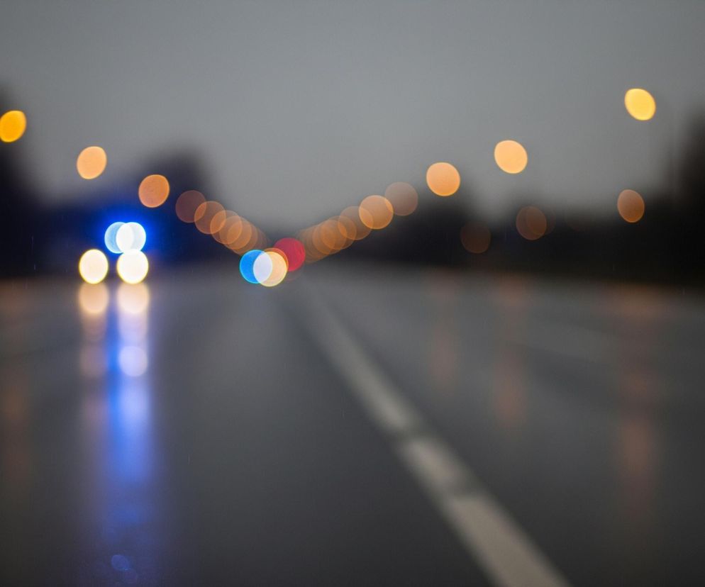 Rozmyty widok ulicy nocą, z drogą biegnącą centralnie ku górze. Asfalt jest ciemny i błyszczący, odbijając światła. Pośrodku drogi widoczna jest przerywana, biała linia. W tle, po obu stronach ulicy, znajdują się liczne rozmyte światła w kolorach pomarańczowym, niebieskim i czerwonym. Po lewej stronie widać wyraźne niebieskie światło i kilka żółtych, natomiast w oddali dominują pomarańczowe kule światła.