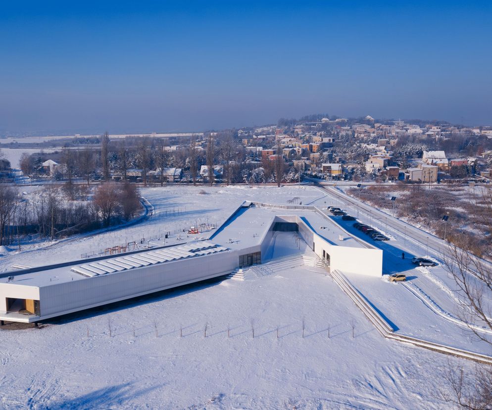 Przedszkole „Ptasie Radio” w Bobrownikach zimą, widok z lotu ptaka. Obiekt o białych elewacjach i płaskich dachach, pokryty śniegiem, otoczony zaśnieżonym terenem. W tle widoczne zabudowania mieszkalne i drzewa. Więcej o architekturze na Architektura Murator Plus.