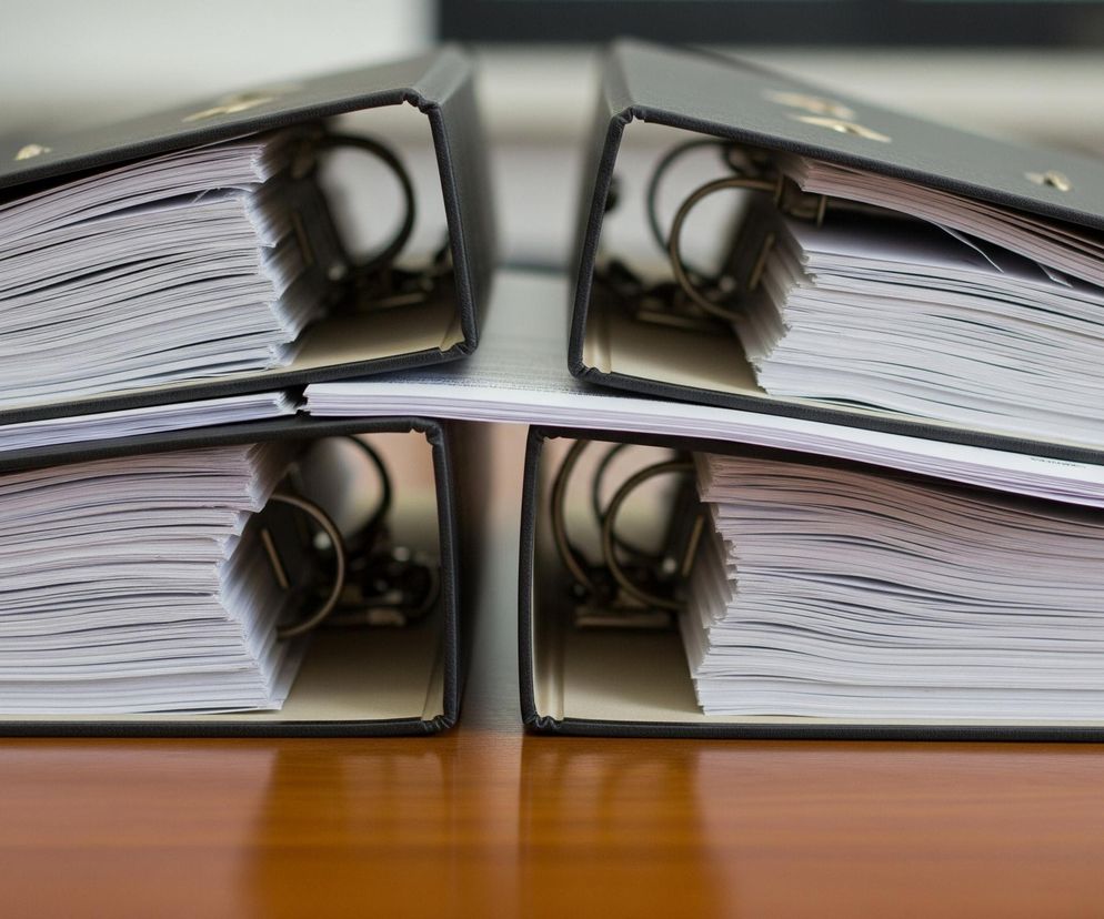 Cztery czarne segregatory z dokumentami stoją pionowo na lśniącej, brązowej powierzchni. Każdy segregator jest wypełniony dużą ilością białych kartek papieru, wystających poza krawędzie okładek. Metalowe, otwarte mechanizmy ringowe są widoczne wewnątrz segregatorów. Tło jest rozmyte, z jasnoszarymi i ciemnymi tonami.