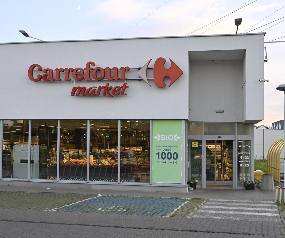Elewacja sklepu Carrefour Market z czerwoną nazwą i strzałką, widoczna w tle domy szeregowe. Przed wejściem szyby witrynowe, a na nich zielone plakaty z logo „BIO” oraz informacją o dostępności ponad 1000 produktów BIO. Obok wejścia regulamin z godzinami otwarcia. W artykule Super Biznes przeczytasz o zakazie handlu.
