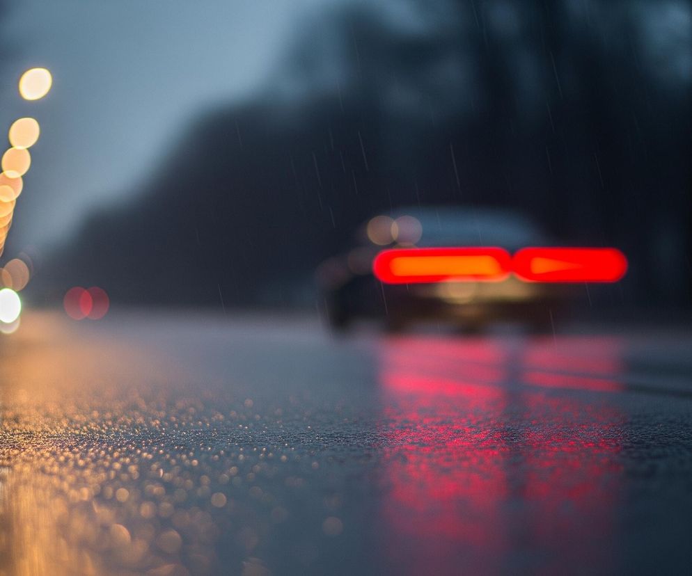 Mokra nawierzchnia drogi w centrum kadru odbija rozmyte światła otoczenia. W oddali, po prawej stronie, widać niewyraźny zarys samochodu z intensywnie czerwonymi światłami tylnymi, które tworzą długie poziome pasy odbite na mokrej powierzchni. Po lewej stronie, wzdłuż krawędzi drogi, ciągnie się rząd rozmytych, pomarańczowych świateł ulicznych, natomiast bliżej, w lewym dolnym rogu, znajdują się dwa białe, okrągłe światła, również odbite w kałużach. Całość utrzymana jest w ciemnej, deszczowej tonacji, z widocznymi drobnymi smugami padającego deszczu.