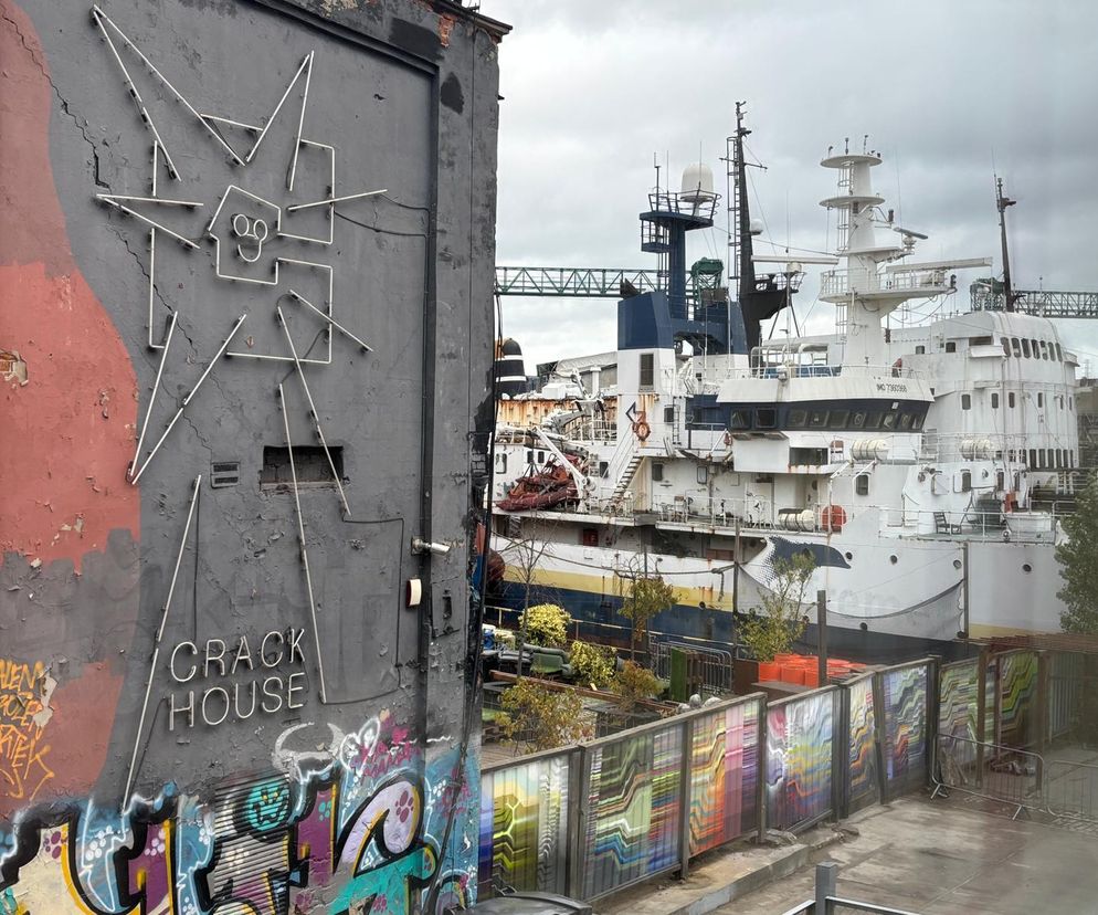 Fragment ściany budynku ze zniszczoną elewacją, pokrytej graffiti i neonową instalacją CRACK HOUSE oraz statkiem badawczym Stoczni Gdańskiej w tle. Ukazuje to przekształcenia terenów poprzemysłowych Gdańska, o których przeczytasz na Architektura Murator Plus.