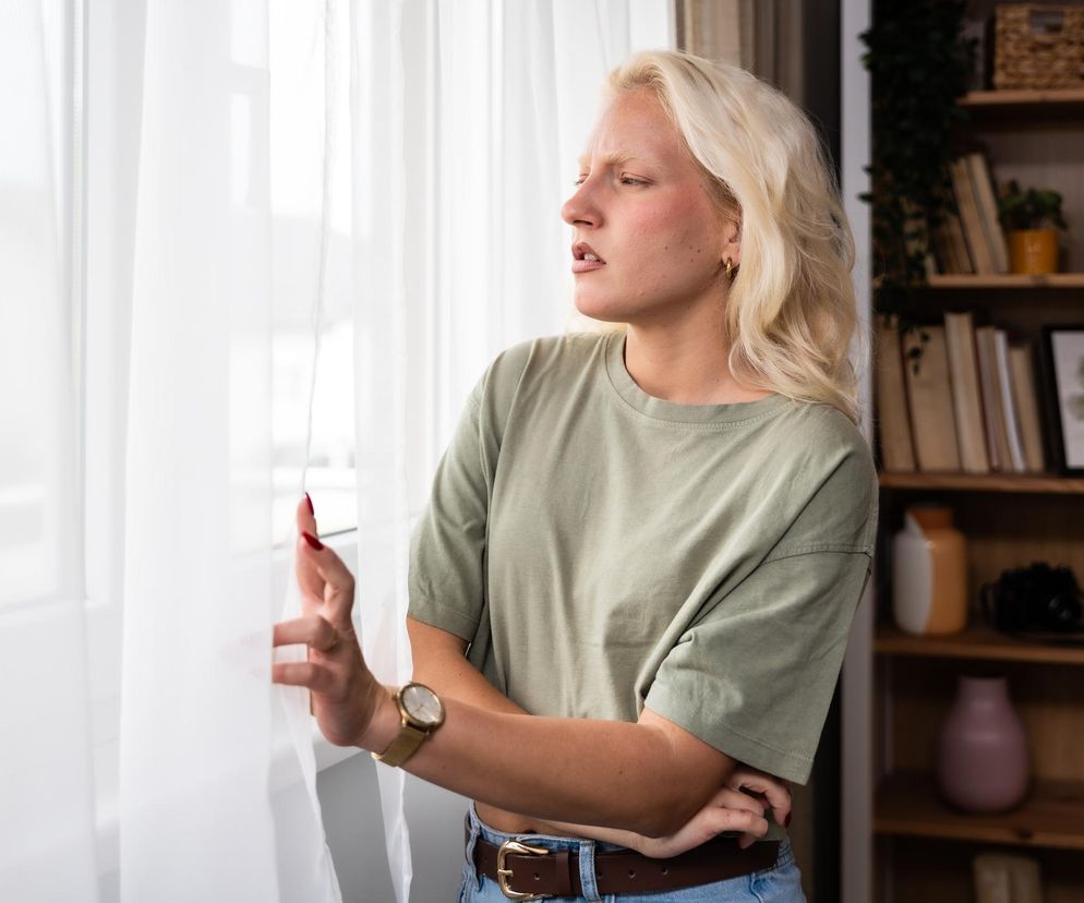 Blondynka o jasnej cerze z marsową miną stoi przy oknie, zerkając w bok, próbując odgrodzić się od hałasu, który niszczy serce i psychikę. To idealna ilustracja problemu poruszonego na portalu Poradnik Zdrowie. Kobieta ubrana jest w zieloną koszulkę i jeansy.