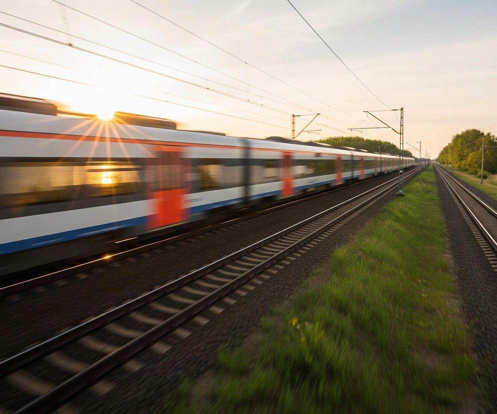 Pędzący pociąg o biało-niebiesko-czerwonym malowaniu zajmuje większą część lewej strony obrazu, rozmazany w ruchu, z promieniami słońca widocznymi w jego oknach. Pociąg porusza się po torach, które biegną równolegle przez cały kadr, rozchodząc się w oddali. Po prawej stronie torów rozciąga się zielony pas trawy i drzew, za którymi widać fragment zbiornika wodnego, a nad wszystkim góruje jasne niebo w odcieniach zachodzącego słońca, poprzecinane cienkimi liniami trakcji.