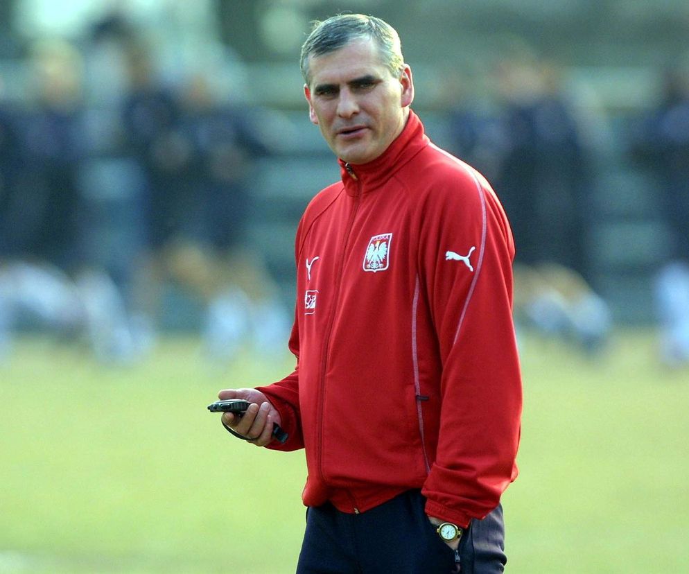 Trener Paweł Janas w czerwonym dresie reprezentacji Polski z orzełkiem, stojący na boisku piłkarskim, trzymający w dłoni telefon komórkowy. Więcej o jego karierze piłkarskiej i trenerskiej przeczytasz na naszym portalu.