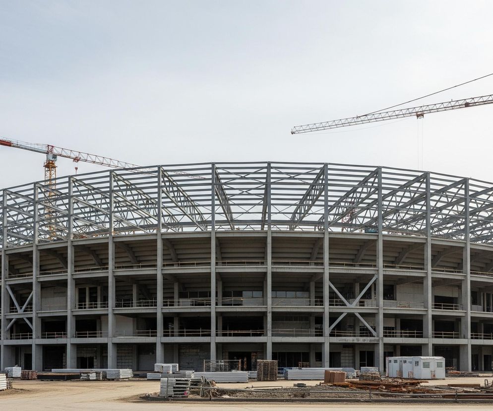 Na pierwszym planie widoczna jest duża, okrągła konstrukcja w trakcie budowy, składająca się z wielu poziomów betonowych filarów i stropów, tworzących otwarte przestrzenie. Górna część konstrukcji to metalowy szkielet dachu. Teren wokół budynku jest pokryty ziemią i gruzem, z licznymi paletami materiałów budowlanych oraz niewielkimi kontenerami budowlanymi. W tle, ponad konstrukcją, wznoszą się trzy wysokie żurawie budowlane o pomarańczowo-żółtych i czerwonych elementach, wskazujące na aktywność budowlaną, pod bezchmurnym, jasnym niebem.