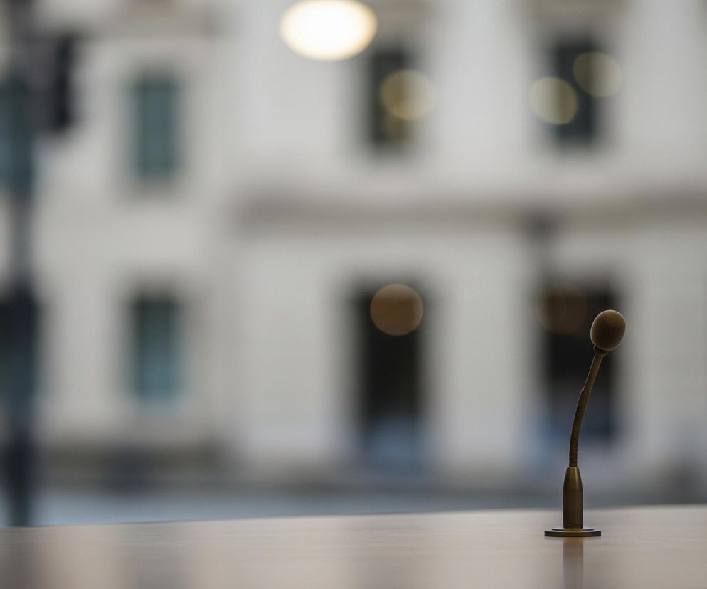 Na pierwszym planie, z prawej strony, znajduje się mały, brązowy mikrofon na ruchomym wysięgniku, osadzony na płaskiej, okrągłej podstawie. Mikrofon umieszczony jest na jasnej, drewnianej powierzchni stołu, która zajmuje dolną część kadru. Tło jest rozmyte, przedstawiając białą fasadę budynku z wieloma ciemnymi oknami oraz jasnożółte, owalne światło u góry po lewej stronie. Lewa krawędź kadru jest częściowo zasłonięta pionową, ciemną kolumną, która rozciąga się od góry do dołu.