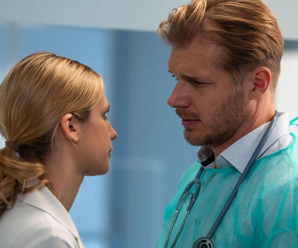 Lekarka Gloria Krasucka i prof. Maks Beger, oboje ubrani w stroje medyczne, patrzą na siebie z powagą w szpitalu w Leśnej Górze. Emocje między nimi rosną. Więcej o ich relacji można przeczytać na Super Seriale.