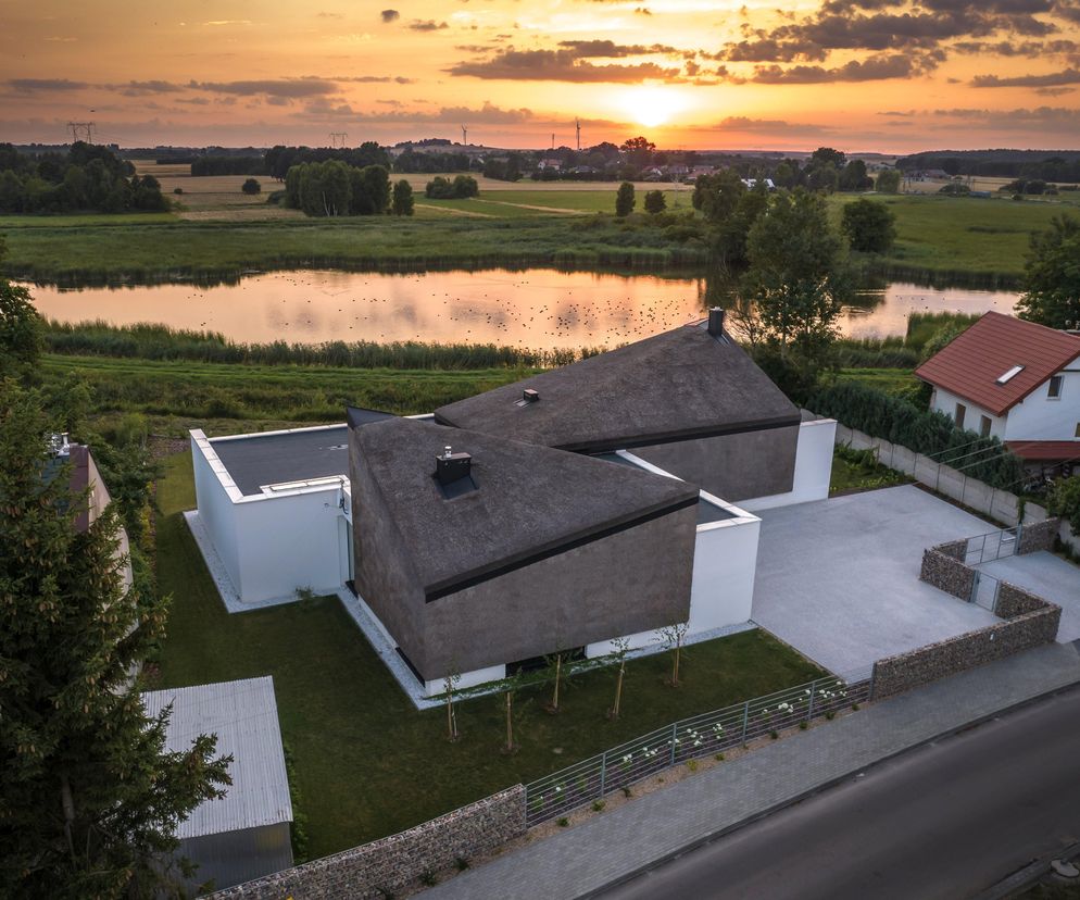 Nowoczesny dom jednorodzinny Barka Cieślika z dachem krytym strzechą, położony nad jeziorem w Białej koło Częstochowy, autorstwa Daniela Cieślika. Widok z lotu ptaka o zachodzie słońca, podkreślający integrację budynku z naturą. O projekcie przeczytasz na Architektura Murator Plus.