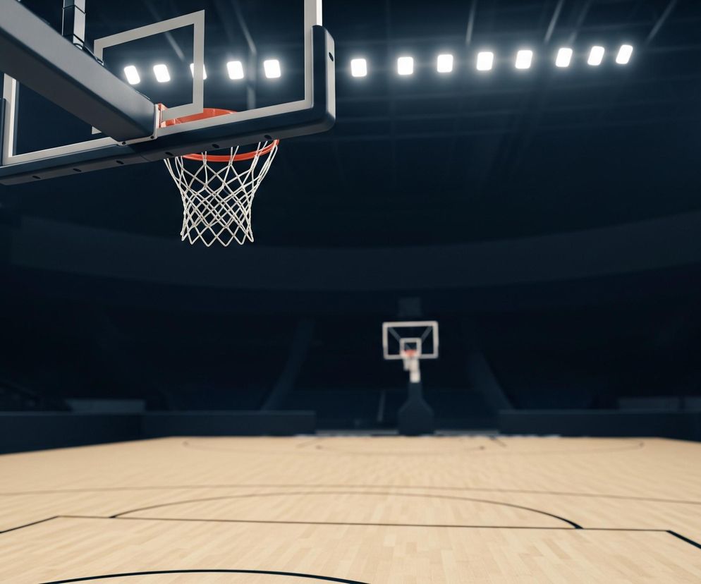 Na pierwszym planie, z góry, widoczna jest obręcz do koszykówki z białą siatką i pomarańczową obwódką, wsparta na czarno-białej konstrukcji. Pod nią rozciąga się boisko o jasnej, drewnianej nawierzchni z czarnymi liniami, które stopniowo zanika w oddali, ukazując kolejną, rozmytą obręcz. W tle, po obu stronach kadru, znajduje się ciemne audytorium z rzędami siedzeń, otoczone przez rozmieszczone równomiernie, kwadratowe reflektory emitujące jasne, białe światło.