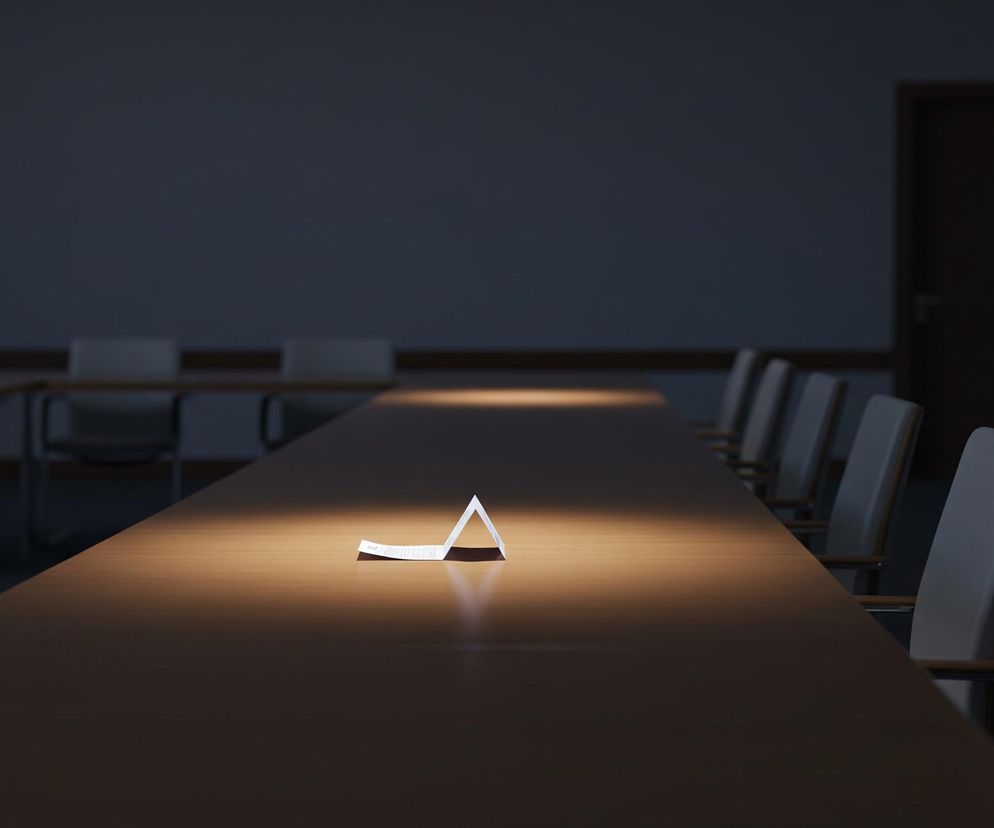 Na ciemnym tle sali konferencyjnej, w centrum uwagi znajduje się długi, brązowy stół. Pośrodku stołu, w miejscu oświetlonym jasnym, ciepłym światłem, leży złożona na kształt namiotu biała kartka papieru, na której widać niewyraźny tekst. Po obu stronach stołu ustawione są rzędy krzeseł z jasnymi, prawdopodobnie szarymi oparciami, częściowo pogrążone w cieniu. W tle, po prawej stronie, widoczne są ciemne, drewniane drzwi.