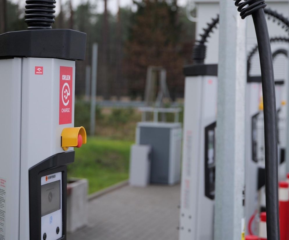 Szereg białych stacji ładowania Orlen Charge, z czerwonymi logo i przyciskami awaryjnymi, na tle lasu. Infrastruktura szybkiego ładowania aut elektrycznych, o której więcej przeczytasz na portalu Super Biznes.