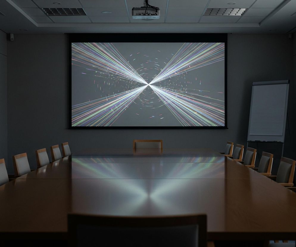 W centrum obrazu znajduje się duży ekran projekcyjny wyświetlający dynamiczną, abstrakcyjną grafikę z wielokolorowymi liniami zbiegającymi się do centralnego punktu na szarym tle, otoczony czarną ramką. Poniżej ekranu rozciąga się długi, drewniany stół konferencyjny w kształcie litery U, o lśniącej powierzchni odbijającej światło. Wokół stołu ustawione są liczne krzesła z jasnymi oparciami i ciemnymi siedziskami. Pomieszczenie ma szare ściany, a na suficie widoczne są kratownice wentylacyjne i projektor zawieszony nad stołem. Po prawej stronie ekranu widać białą tablicę na sztalugach, a po lewej brązowe drzwi z metalową klamką, nad którymi znajduje się zielony piktogram wyjścia ewakuacyjnego.