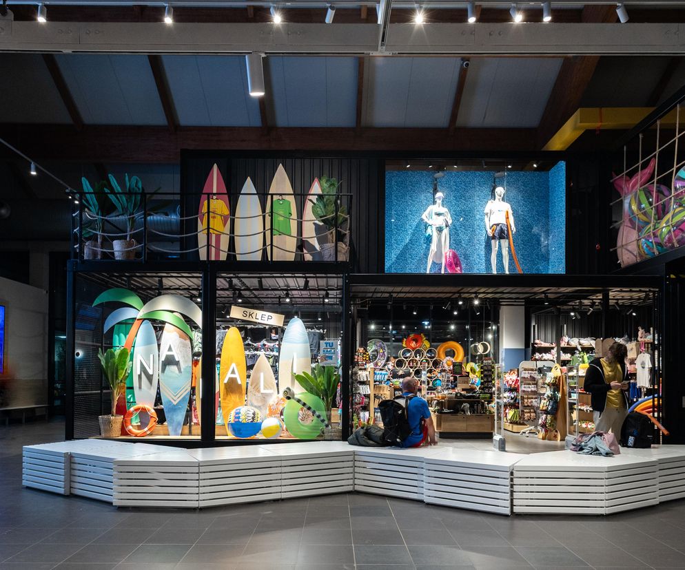 Nowoczesny hol wejściowy w Aquaparku Fala, projekt RWSL, z elementami nawiązującymi do nadmorskich wakacji i portowego wybrzeża. Widać strefę sklepową z manekinami i akcesoriami pływackimi, oraz wygodne, białe ławki. O tej aranżacji czytaj więcej na Architektura Murator Plus.