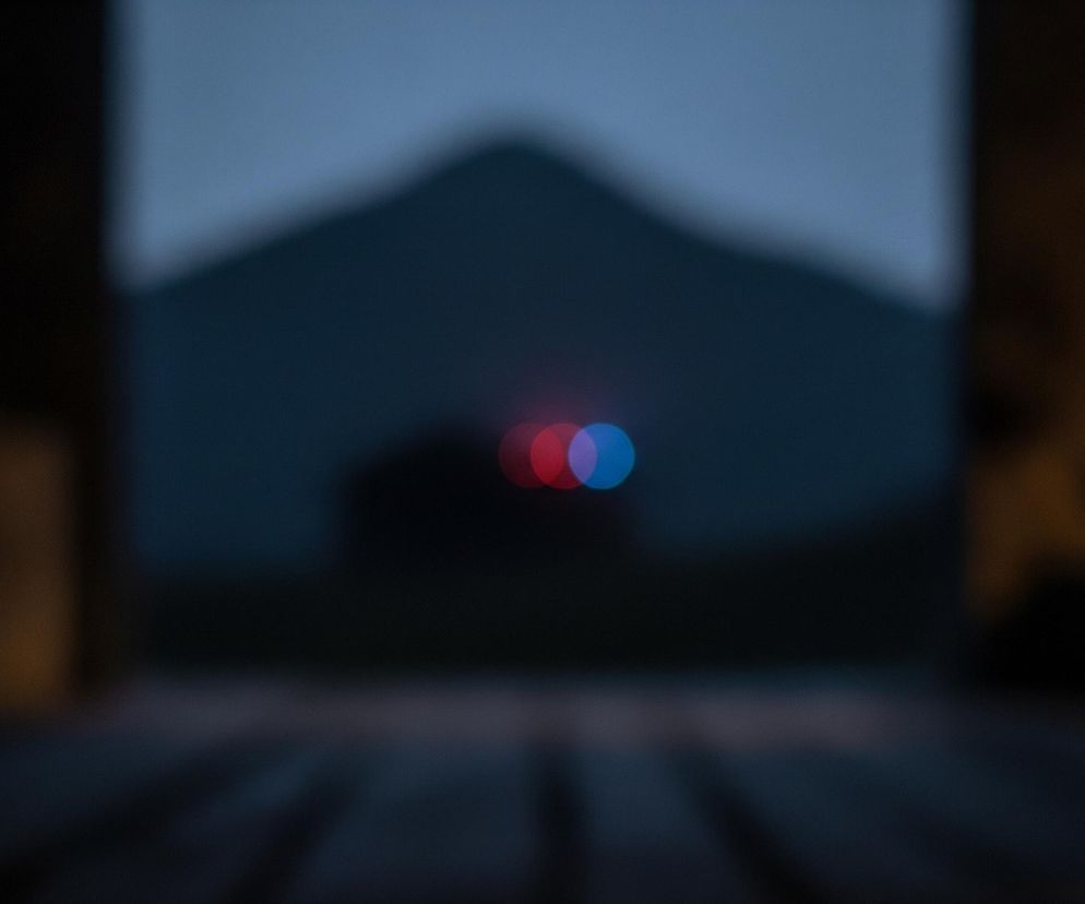 Centralnym punktem obrazu są dwa rozmyte, koliste światła w odcieniach czerwieni i błękitu, umieszczone na tle nieostrego, ciemnego krajobrazu. Tło ukazuje zarys góry lub wzniesienia o ciemnoniebieskim odcieniu, które kontrastuje z jaśniejszym, rozmytym niebem. Cała scena jest obserwowana przez ciemną ramę, prawdopodobnie bramy lub okna, z widocznymi nieostrymi drewnianymi deskami lub belkami, które tworzą dolną i boczne krawędzie obrazu.