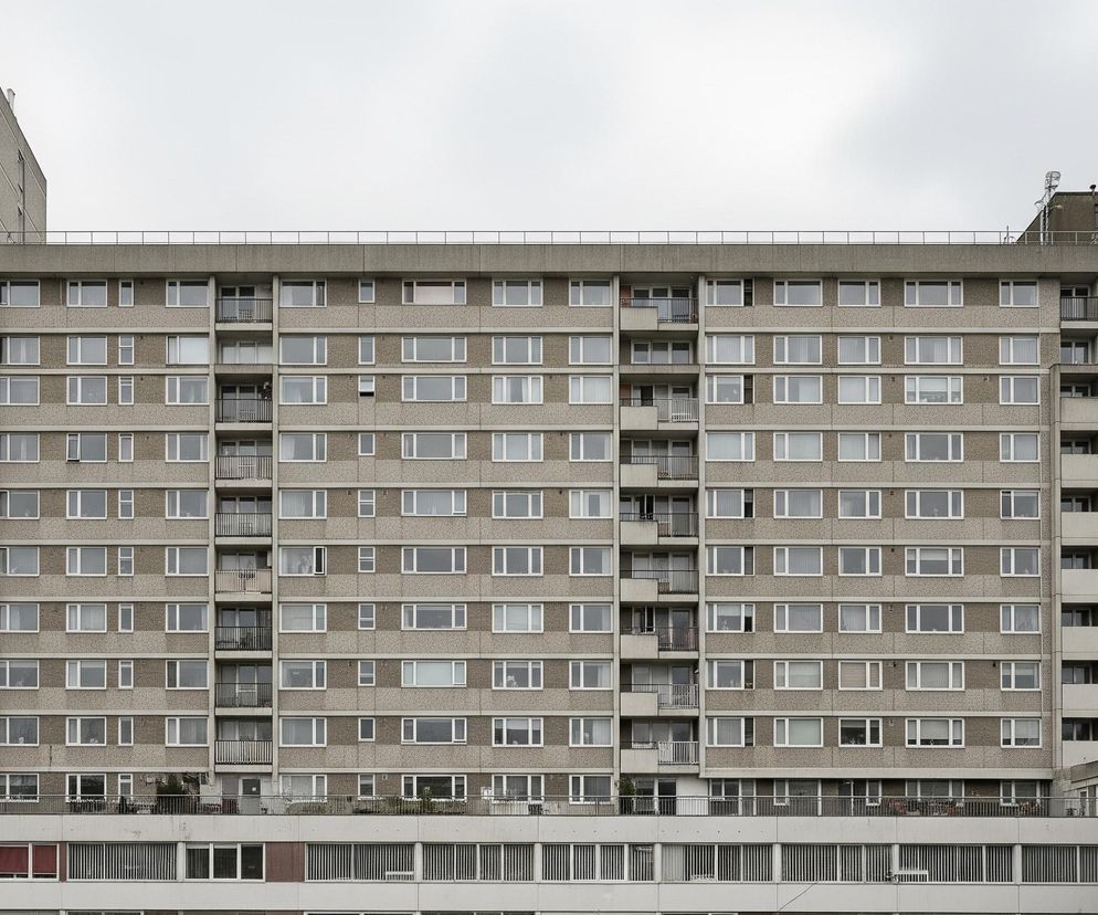 Dominującym elementem jest wysoki, prostokątny budynek mieszkalny o szarej, betonowej fasadzie, pokrytej drobnym, ziarnistym wzorem. Liczne okna o białych ramach są rozmieszczone regularnie na całej szerokości budynku, a pomiędzy nimi znajdują się poziome pasy gładkiego betonu oraz pionowe, wąskie balkony z czarnymi, metalowymi balustradami. Po lewej stronie fasada budynku jest jednolita, a po prawej widoczny jest niższy fragment, również pokryty oknami, połączony z główną bryłą. Pod pochmurnym, jasnoszarym niebem, po lewej stronie u dołu, częściowo zasłonięta, stoi szara latarnia uliczna.