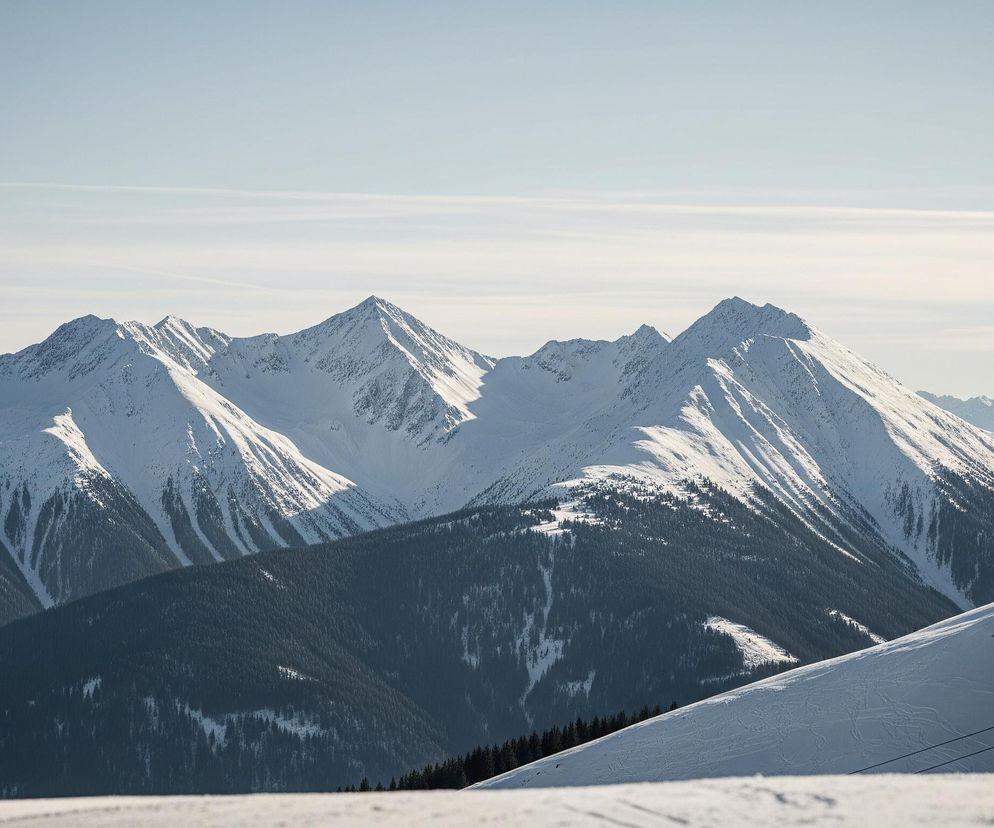 Góry pokryte śniegiem dominują krajobraz, z wyraźnymi grzbietami i zboczami oświetlonymi słońcem, kontrastującymi z cienistymi fragmentami. W dolnych partiach gór rośnie ciemnozielony las iglasty. Na pierwszym planie, w prawym dolnym rogu, widoczny jest fragment stoku narciarskiego z zarysami tras i pojedynczym krzesełkiem wyciągu narciarskiego. Niebo jest jasnoniebieskie, z delikatnymi smugami chmur i pojedynczym białym smugowym śladem za samolotem.