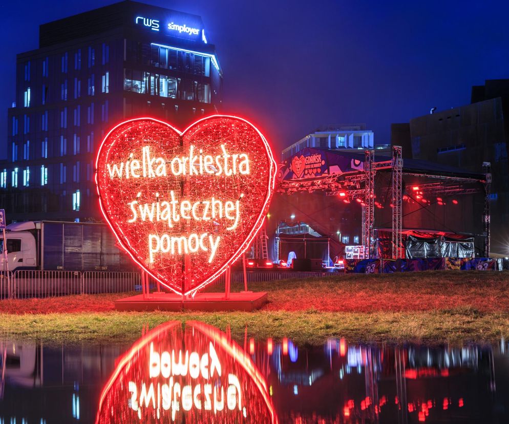 Duże, czerwone neonowe serce z napisem Wielka Orkiestra Świątecznej Pomocy stoi na tle nocnego nieba, odbijając się w wodzie. W tle widoczny jest budynek biurowy oraz scena z konstrukcją oświetleniową, co ilustruje przygotowania do Finału WOŚP, o którym można przeczytać na portalu Poradnik Zdrowie.