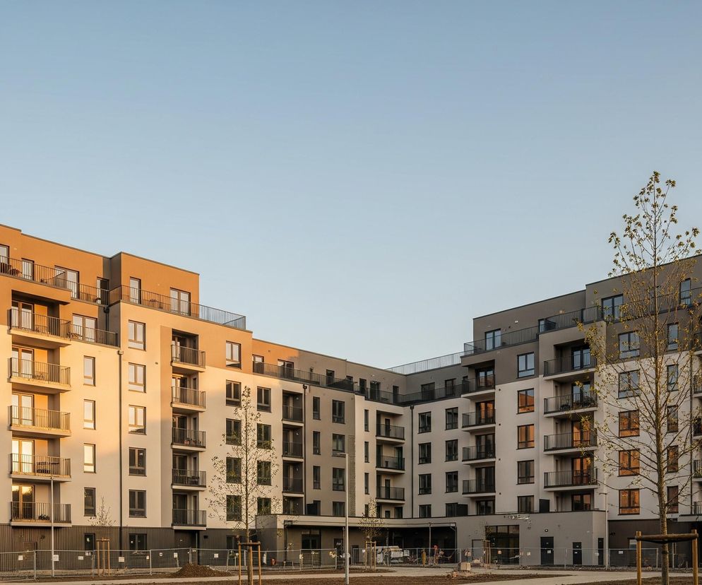 Przedstawiono kompleks mieszkalny o nowoczesnej architekturze, składający się z kilku połączonych budynków, rozmieszczonych wokół otwartego dziedzińca. Lewa strona budynków jest oświetlona ciepłym, złotym światłem zachodzącego słońca, podkreślającym pomarańczowo-brązowe i beżowe fasady, z licznymi oknami i balkonami z ciemnymi balustradami. Prawa część kompleksu pozostaje w cieniu, ukazując szare i ciemnoszare elewacje z analogicznym układem okien i balkonów. Na pierwszym planie widoczne są młode drzewa z drobnymi liśćmi, ogrodzone tymczasowymi siatkami, oraz niezabudowany teren z widoczną ziemią i fragmentami wybrukowanej powierzchni. Bezchmurne, jasnoniebieskie niebo stanowi górną część obrazu.