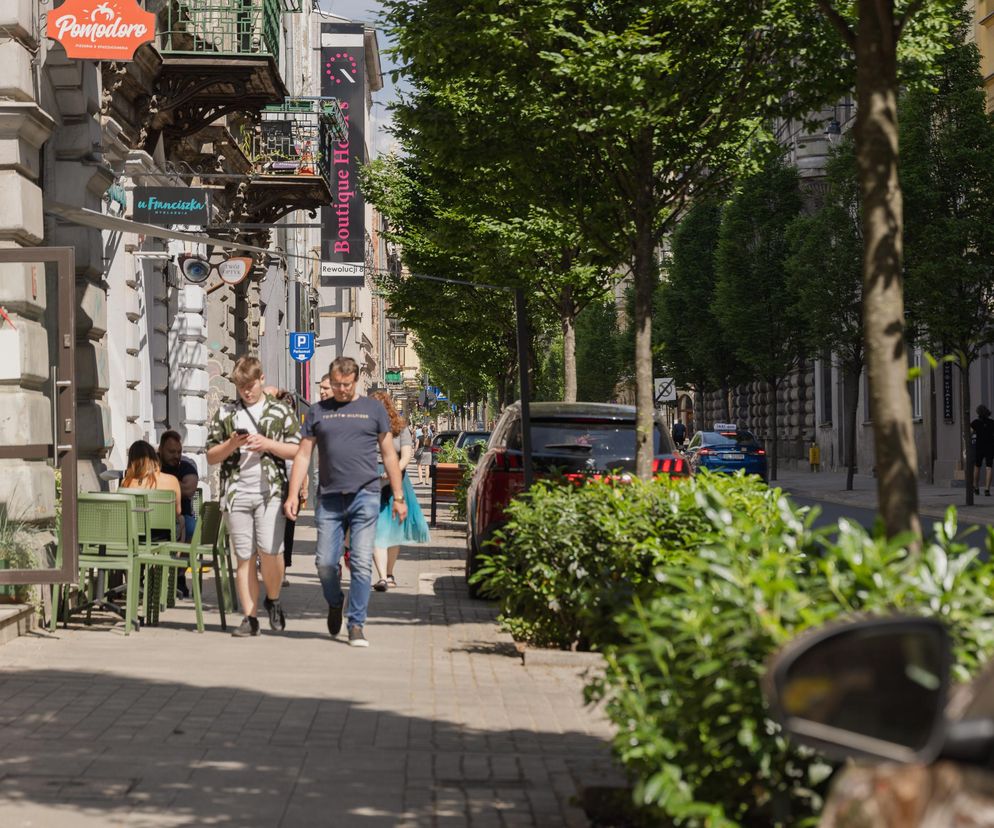 Ulica Wschodnia w Łodzi, pełna zieleni i tętniąca życiem, z widocznymi spacerującymi ludźmi, kawiarnianymi ogródkami oraz budynkami z pięknymi elewacjami. To przykład rewitalizacji miejskiej przestrzeni, o której można przeczytać na portalu Architektura Murator Plus.