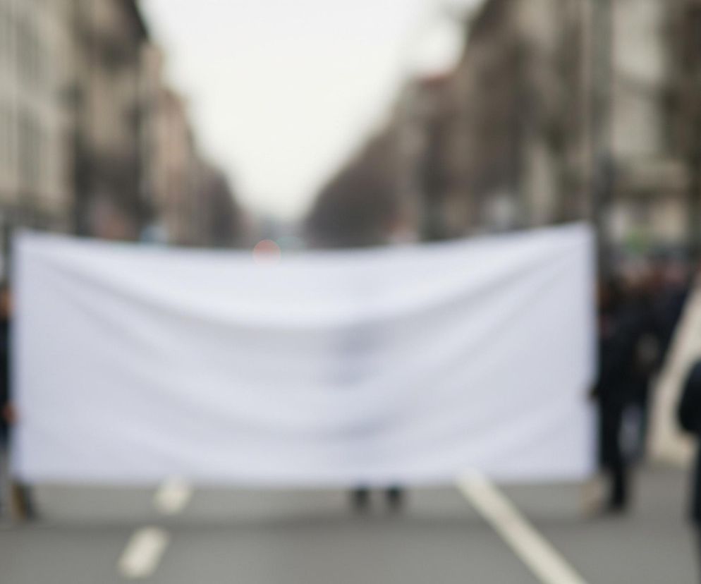 Zamazana scena uliczna przedstawiająca grupę osób niosących duży, biały transparent. Transparent zajmuje centralną część kadru, jest rozciągnięty poziomo i zakrywa sporą część tła. Osoby go niosące oraz pozostałe postacie na ulicy są rozmyte i nieostre. Tło tworzą zamazane fasady budynków po obu stronach ulicy oraz szare niebo nad nimi. Na asfalcie widoczne są niewyraźne, jasne linie poziome.