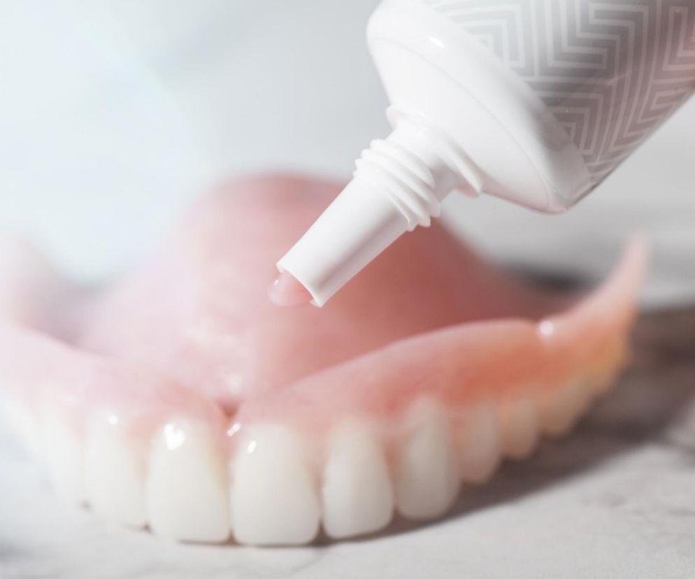 Proteza zębowa z różowymi dziąsłami, do której osoba wyciska specjalny klej lub pastę mocującą z białej tubki. Obraz symbolizuje prawidłową pielęgnację i codzienne użytkowanie protezy, o czym przeczytasz więcej na Poradnik Zdrowie.