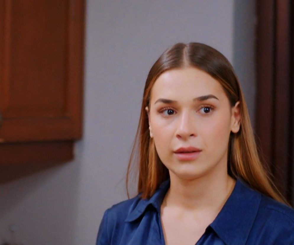 Aktorka Sila z serialu Miłość i nadzieja w niebieskiej koszuli z kołnierzykiem, w półzbliżeniu na twarz, wyrażająca zmartwienie, stojąca w pomieszczeniu. W tle widoczne fragmenty brązowej szafki i jasnej ściany. Więcej o losach bohaterów przeczytasz na Super Seriale.