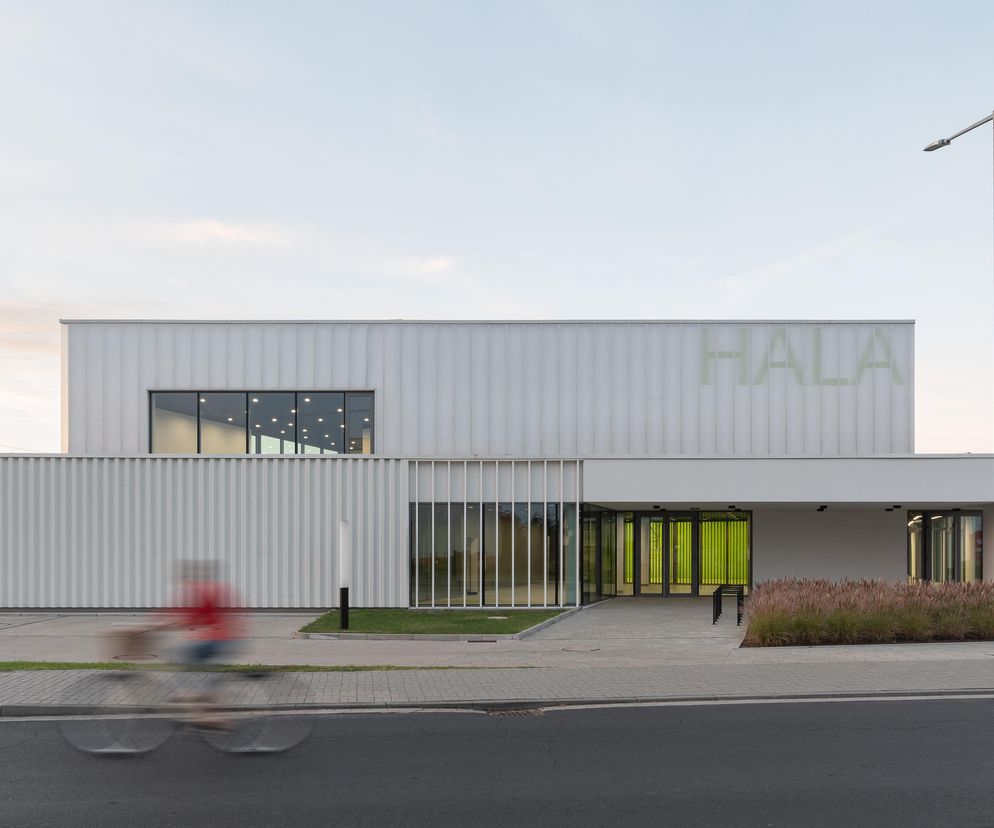 Jasnoszara hala widowiskowo-sportowa w Książu Wielkopolskim, projekt PL.ARCHITEKCI, Bartłomieja Bajona i Katarzyny Cynki-Bajon. Fasada z blachy trapezowej i poliwęglanu, z podświetlonym wejściem w kolorze limonki i napisem H/A/L/A/ na górze. Po lewej stronie niewyraźna postać na rowerze. O tej realizacji przeczytasz więcej na Architektura Murator Plus.