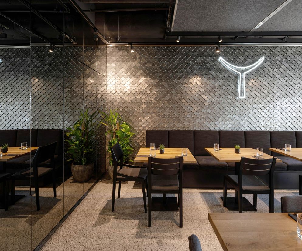 Wnętrze restauracji rybnej Tuna z unikalnymi ścianami pokrytymi niemal 25 tysiącami pokrywek od puszek, imitujących rybie łuski. Na zdjęciu widać drewniane stoły, ciemne krzesła i kanapy. Neonowe logo restauracji podkreśla morski styl, o którym można przeczytać na Architektura Murator Plus.
