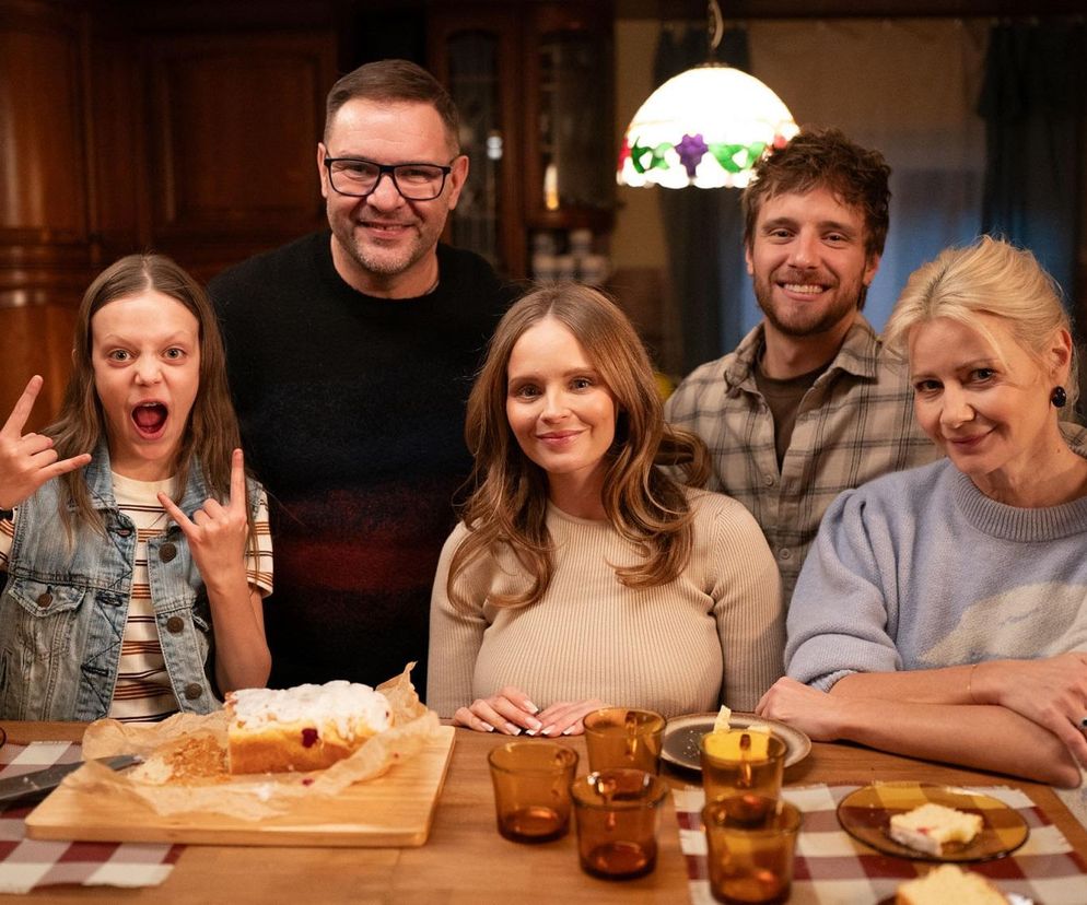 Aktorzy serialu Rodzinka.pl Małgorzata Kożuchowska (Natalia), Tomasz Karolak (Ludwik), Adam Zdrójkowski (Kuba) oraz dwie młode kobiety pozują razem przy stole, na którym leży ciasto. Wszyscy uśmiechają się do kamery, co symbolizuje rodzinne chwile. Więcej o kryzysie w rodzinie Boskich przeczytasz na Super Seriale.