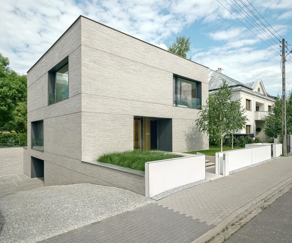 Nowoczesny Dom Rozcięty autorstwa PL.ARCHITEKCI, zgłoszony do konkursu Życie w Architekturze 2025. Prosta bryła z szarej cegły, duże okna, minimalistyczne detale, zjazd do garażu, roślinność i białe murki. Architektura Murator Plus prezentuje zgłoszenie.