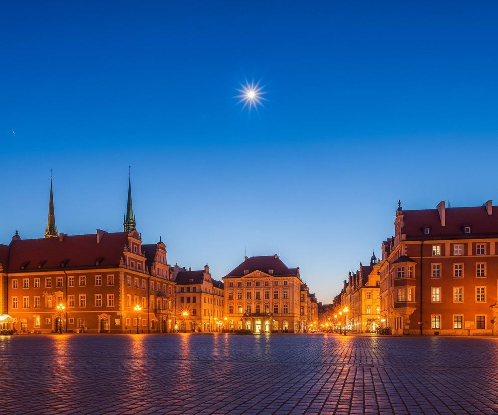 Rozległy plac wybrukowany kostką, odbijającą pomarańczowe światła ulicznych latarni, rozciąga się w centrum kadru, prowadząc wzrok w głąb perspektywy. Otaczają go wielopiętrowe kamienice o zróżnicowanej architekturze, z oświetlonymi oknami i elementami elewacji, niektóre z nich posiadają czerwone dachy i charakterystyczne spiczaste wieże. Nad budynkami rozpościera się ciemnobłękitne, wieczorne niebo, a na nim wyraźnie widoczna jest jasna, rozświetlona gwiazda z promieniami oraz delikatny, wąski sierp księżyca.