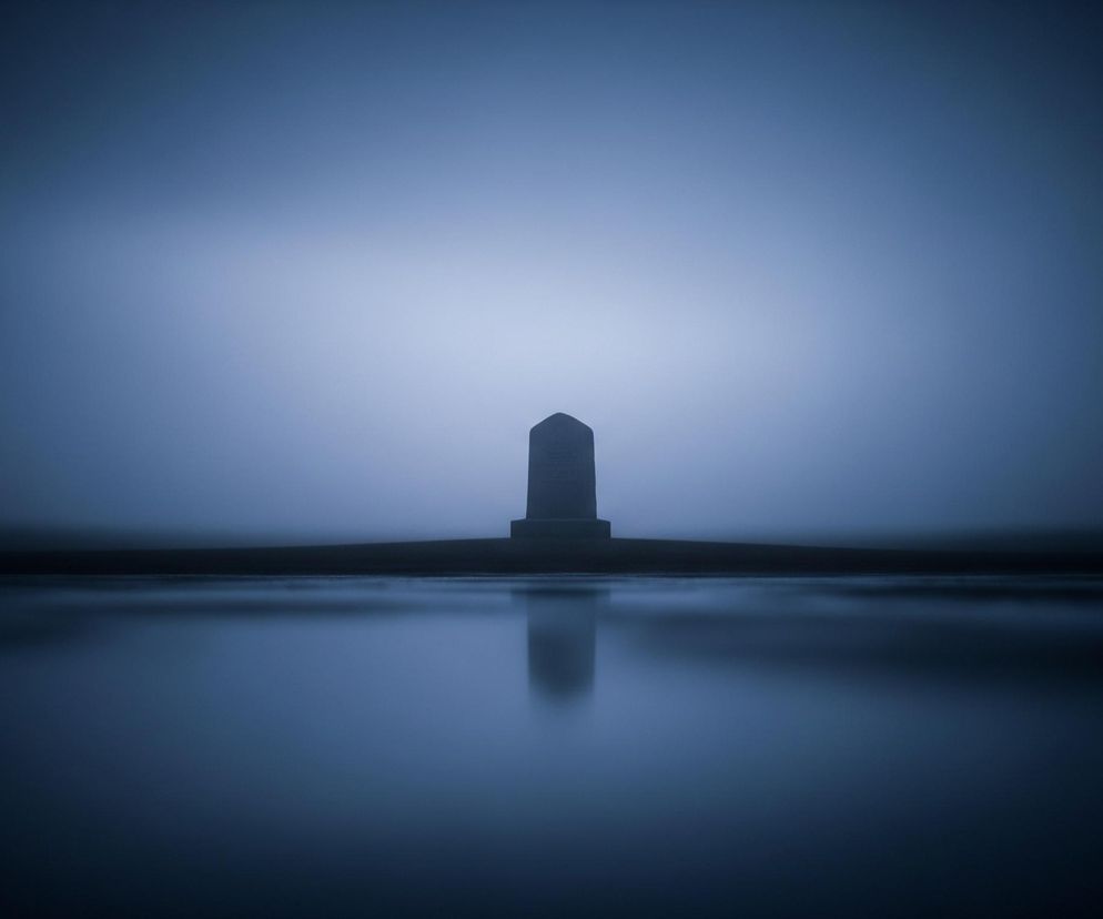 Na zdjęciu centralnie umieszczony jest ciemny, stożkowaty obelisk, zwężający się ku górze i zakończony kopułą. Obiekt stoi na horyzontalnym, ciemnym cokole, rozciągającym się na boki. Poniżej obelisku znajduje się idealnie gładka powierzchnia wody, w której odbija się jego sylwetka oraz pojaśnienie zza obelisku. Tło stanowi zamglone, rozmyte niebo o zmiennym odcieniu błękitu, jaśniejsze w centrum i ciemniejące ku krawędziom. Całość utrzymana jest w jednolitej, chłodnej tonacji granatowo-niebieskiej.