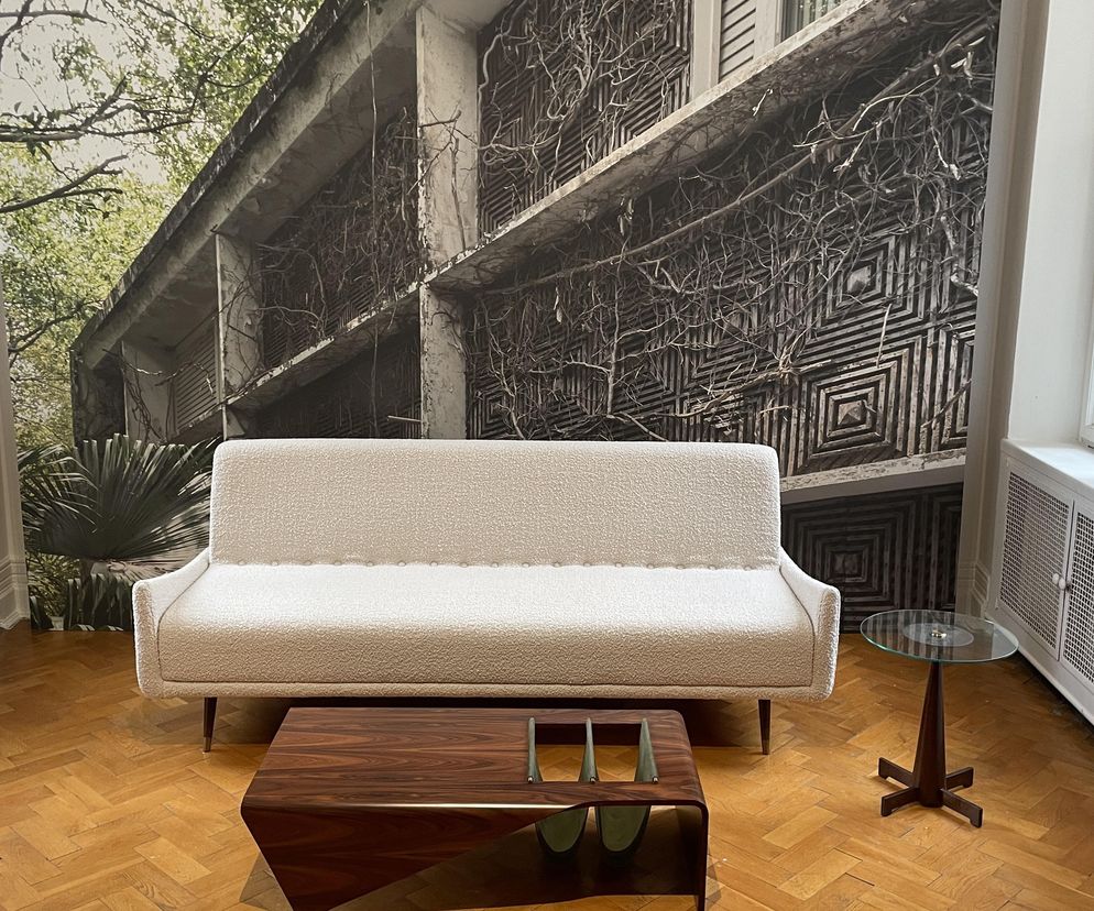 Wnętrze wystawy Warszawa – São Paulo – Warszawa z beżową kanapą i drewnianym stolikiem kawowym w stylu modernizmu Jerzego Zalszupina. Na tle ściany mural przedstawiający zabytkową architekturę z gęstą roślinnością. Otworzyło się dla zwiedzających, o czym przeczytasz na Architektura Murator Plus.