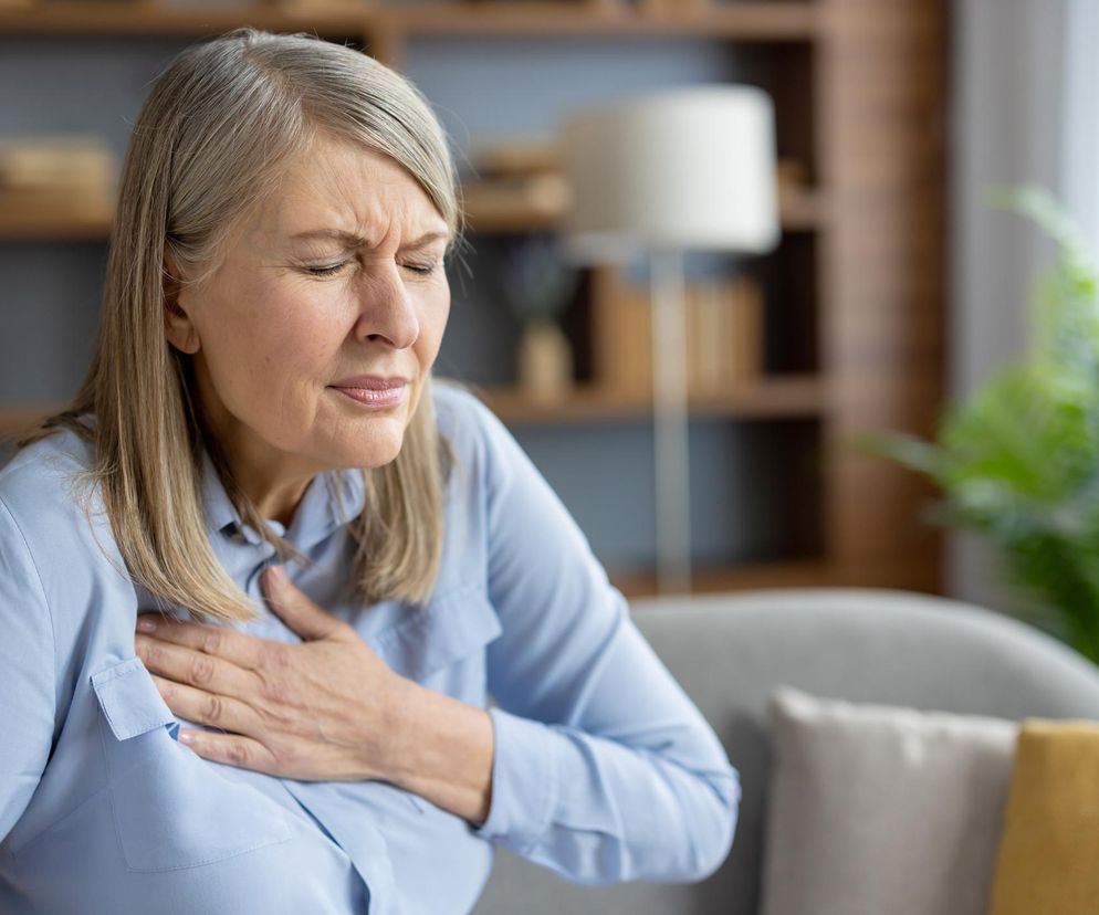 Starsza kobieta w jasnoniebieskiej koszuli, z dłońmi zaciśniętymi na klatce piersiowej, wyraża ból i dyskomfort. Jest to wizualne odzwierciedlenie problemów kardiologicznych, o których możesz przeczytać więcej na portalu Poradnik Zdrowie.