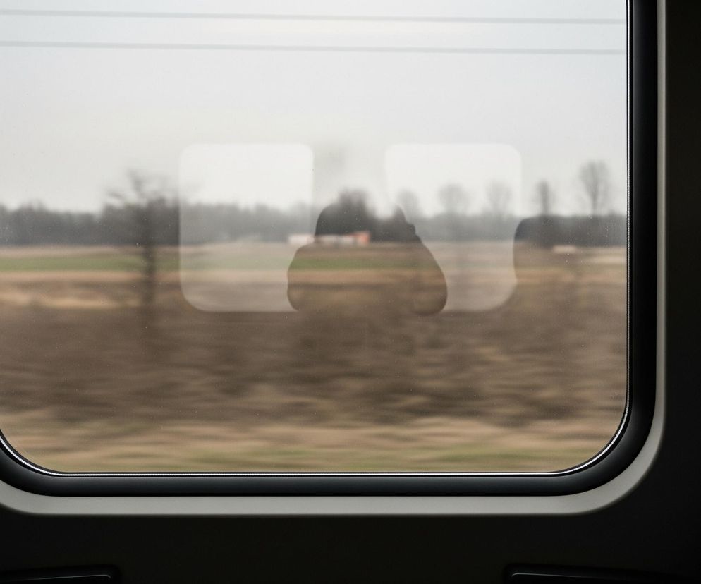 Widok zza okna pociągu przedstawia rozmyty krajobraz z brązowym polem na pierwszym planie i ciemnozielonymi drzewami na horyzoncie, pod szarym niebem. W szybie odbijają się zarys dwóch prostokątnych, rozświetlonych obiektów oraz niewyraźna sylwetka osoby. Wnętrze pociągu widoczne jest po bokach okna, z fragmentami ciemnych siedzeń pokrytych granatową tkaniną w żółte kropki.
