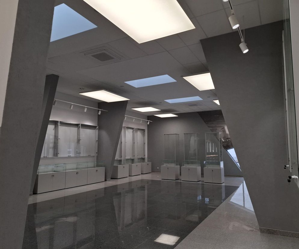 Wnętrze Sali Tradycji Lotniczej Akademii Wojskowej w Dęblinie z przeszklonymi gablotami ekspozycyjnymi i nowoczesnym oświetleniem. Pomieszczenie ma szare ściany i błyszczącą posadzkę, a sufit wyposażono w podłużne świetliki. Więcej o projekcie Pawła Pływacza przeczytasz na Architektura Murator Plus.