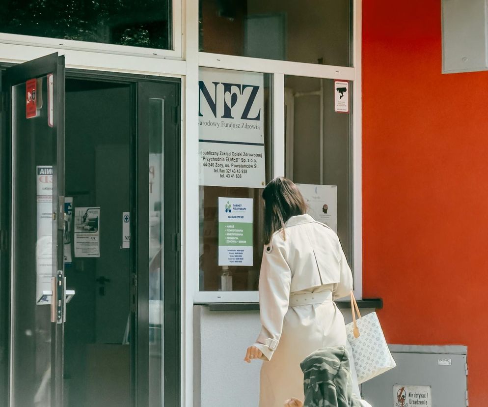 Kobieta w beżowym płaszczu wchodząca do budynku z logo Narodowego Funduszu Zdrowia, symbolizująca kwestię utraty prawa do darmowego leczenia po 30 dniach od ustania ubezpieczenia. Więcej o zmianach w opiece zdrowotnej przeczytasz na Super Biznes.