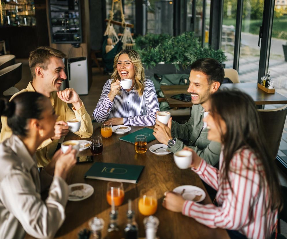 Grupa pięciu roześmianych młodych ludzi siedzi przy drewnianym stole w kawiarni z panoramicznymi oknami, pijąc kawę i soki. Kobieta w centrum z blond włosami w biało-niebieskiej koszuli śmieje się głośno. Poradnik Zdrowie przypomina, że takie spotkania to klucz do szczęścia.