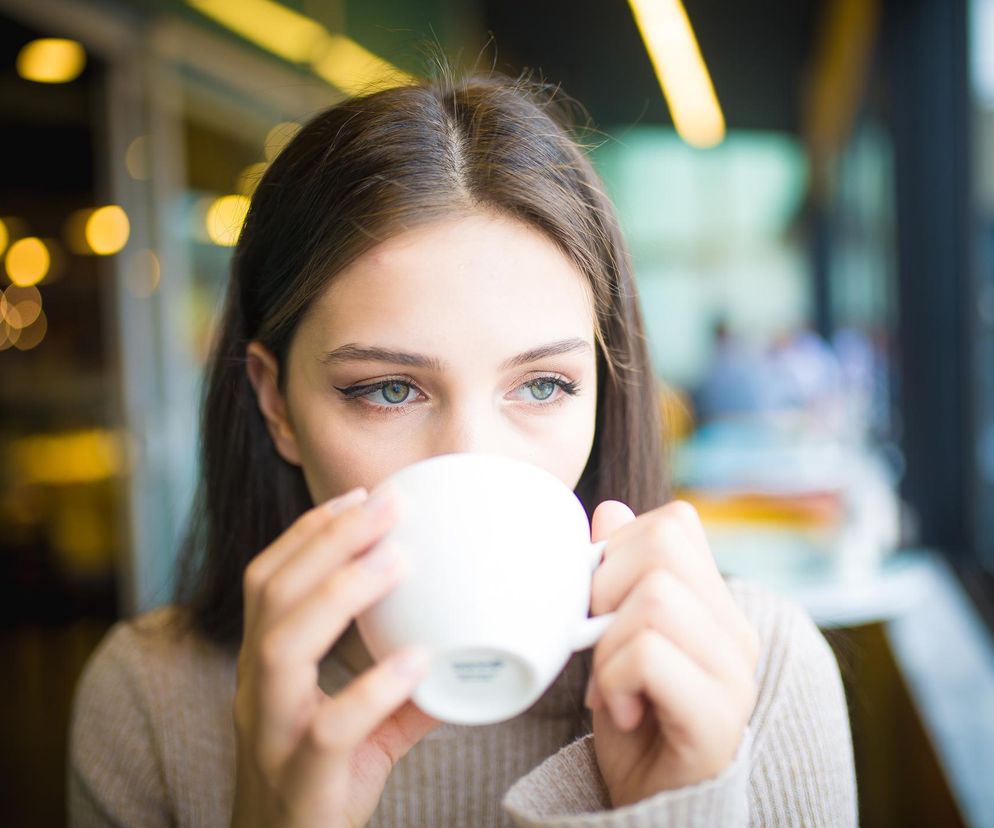 Młoda kobieta z długimi, ciemnymi włosami, w beżowym swetrze, pije kawę z białego kubka, patrząc w bok z delikatnym uśmiechem na twarzy. Tło jest rozmyte, z widocznymi jasnymi punktami i oknem, co sugeruje wnętrze kawiarni. Dowiedz się więcej o zdrowym piciu kawy na Poradnik Zdrowie.